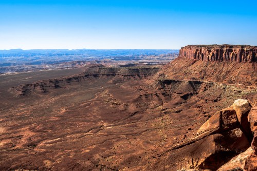 A vast desert landscape with red rock formations and a clear blue sky, showcasing rugged terrain and distant mesas.