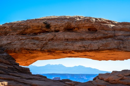 A natural rock arch frames a distant mountain range against a clear blue sky.