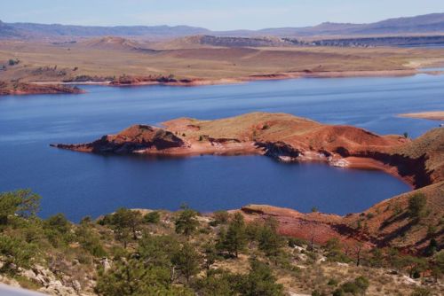 A serene lake surrounded by red rock formations and green trees, with rolling hills in the background under a clear blue sky.