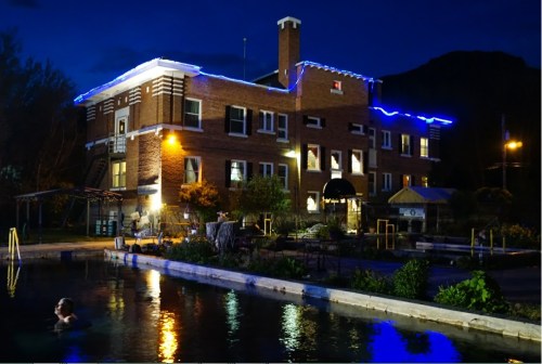 A brick building illuminated with blue lights at night, beside a calm pool reflecting the lights and surrounding nature.
