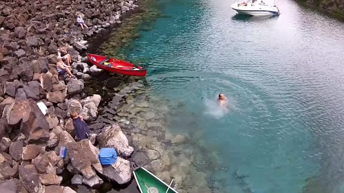 A person swims in clear water near rocky shores, with boats and people relaxing nearby.