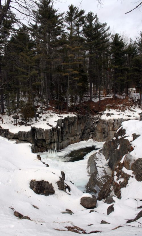 A snowy landscape featuring a rocky cliff and a frozen river, surrounded by tall pine trees.