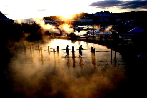 Two people walk through a steaming hot spring at sunset, with mist rising and a scenic landscape in the background.