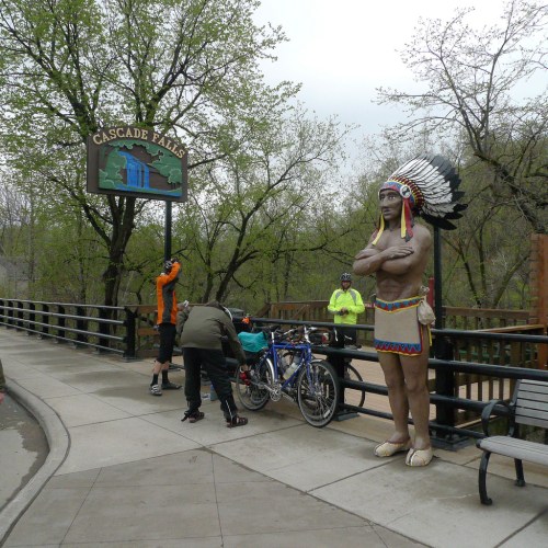 A statue of a Native American with a feathered headdress stands near Cascade Falls, with cyclists nearby.