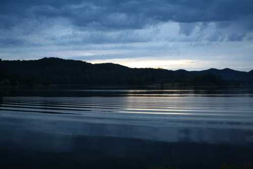 A serene lake at dusk, with gentle ripples and dark mountains silhouetted against a cloudy sky.