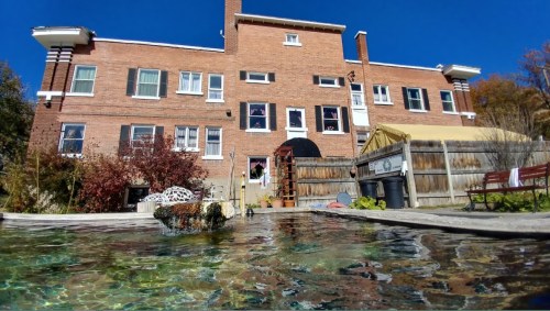 A brick building with multiple windows overlooks a pool, surrounded by greenery and a wooden fence under a clear blue sky.