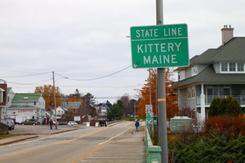 Green state line sign for Kittery, Maine, with a street view of nearby buildings and autumn foliage.