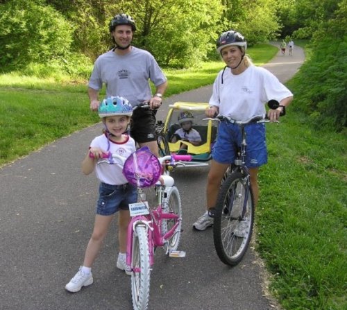 A family biking on a path, with a girl on a pink bike and a toddler in a bike trailer. Green trees in the background.