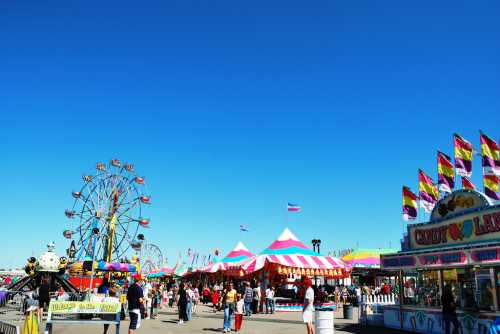 A vibrant carnival scene with colorful tents, a Ferris wheel, and people enjoying various attractions under a clear blue sky.