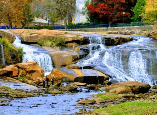 A scenic waterfall cascades over rocks, with two people exploring the riverbank amidst autumn foliage.