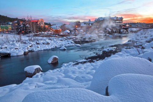 A snowy landscape with a river, steam rising, and buildings in the background under a colorful sunset sky.