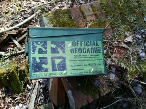 A green geocache container with the text "Official Geocache" on a wooded background.