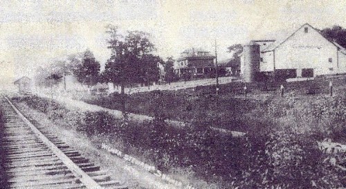 Black and white photo of a railway track with a house and barn in the background, surrounded by trees and greenery.