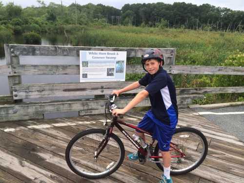 A boy in a helmet poses on a bike near a sign about White Horn Brook and Genosee Swamp, with a wetland backdrop.