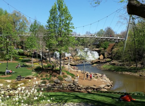 A scenic park with a waterfall, a river, walking bridge, and people enjoying the outdoors on a sunny day.