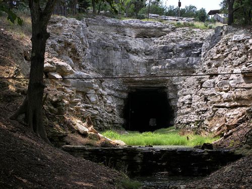 A rocky cave entrance surrounded by greenery, with a small stream in the foreground and trees on either side.