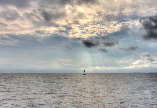 A green buoy stands in calm waters under a cloudy sky with rays of light breaking through the clouds.