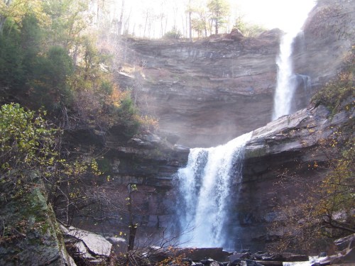 A majestic waterfall cascades down rocky cliffs, surrounded by lush greenery and mist in a serene natural setting.