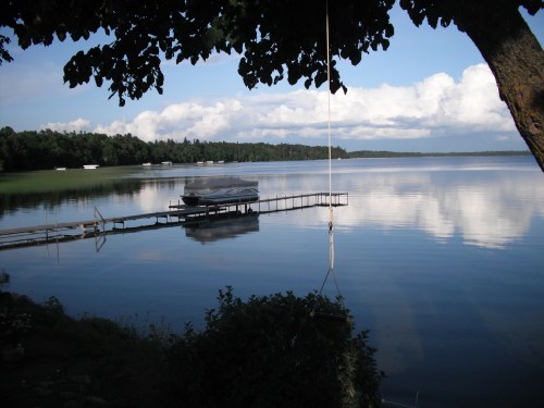 A serene lake scene with a dock, a boat, and reflections of clouds in the water, framed by trees.