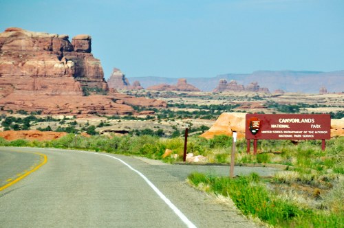 A scenic view of a road leading to the Canyonlands National Park sign, surrounded by red rock formations and desert landscape.