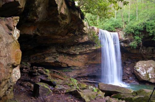 A serene waterfall cascades over rocks into a tranquil pool, surrounded by lush greenery and rugged cliffs.