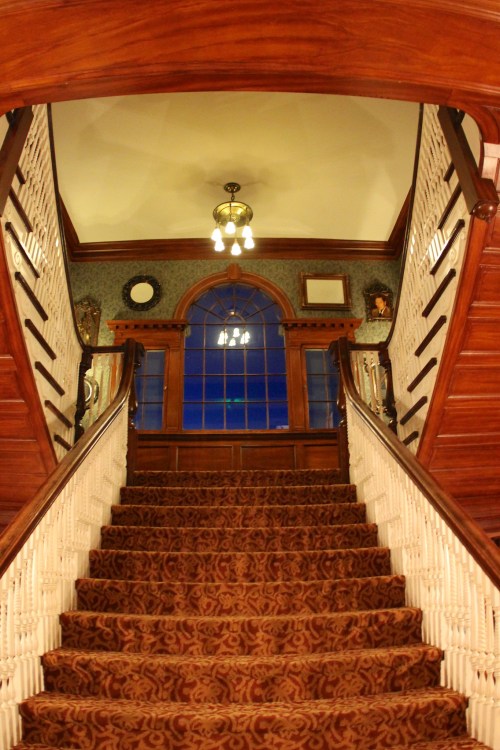 A grand staircase with ornate wooden railings, leading to a windowed landing with vintage decor and warm lighting.