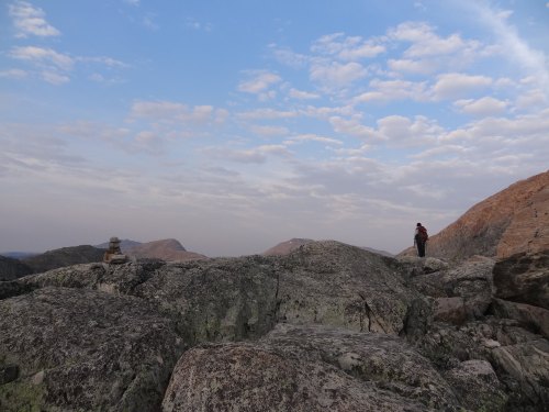 A person stands on rocky terrain under a cloudy sky, with mountains in the background and a small stone stack nearby.