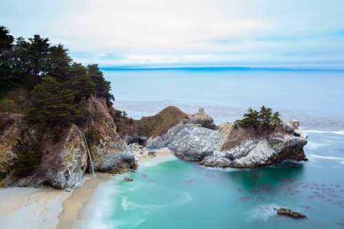 A serene coastal scene featuring a waterfall cascading onto a sandy beach, surrounded by rocky cliffs and lush greenery.