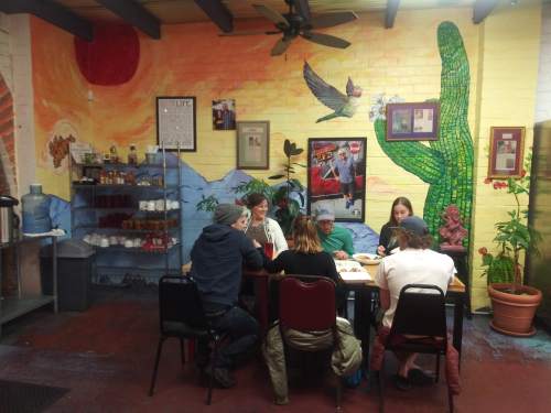 A group of people sits around a table in a colorful restaurant with a mural featuring a sunset and cacti.