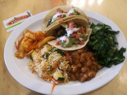 A plate of tacos topped with guacamole and salsa, served with rice, beans, greens, and a side of chips.