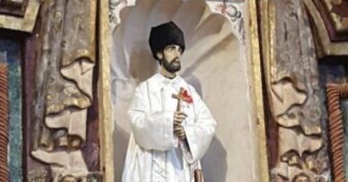 A statue of a man in white robes and a black hat, holding a cross, set in an ornate niche.