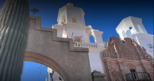 A view of ornate white buildings with towers against a clear blue sky, framed by a cactus and stone archway.
