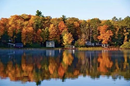 A serene lake reflecting vibrant autumn trees and cozy cabins along the shore under a clear blue sky.