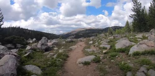 A rocky trail winds through a lush landscape with trees and mountains under a partly cloudy sky.
