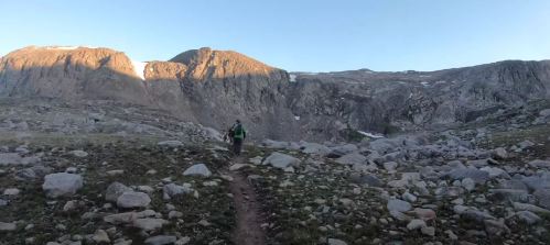 A hiker walks along a rocky trail in a mountainous landscape under a clear blue sky.