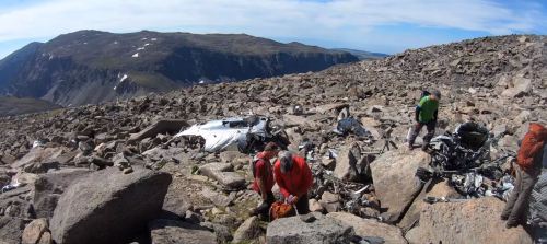 Rescue team members search a rocky landscape for debris from a plane crash, with mountains in the background.