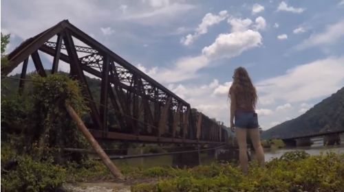 A person stands by a river, gazing at an old, rusted train bridge under a blue sky with scattered clouds.