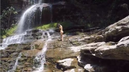 A person stands near a waterfall, surrounded by lush greenery and rocky terrain. Water cascades down the rocks.