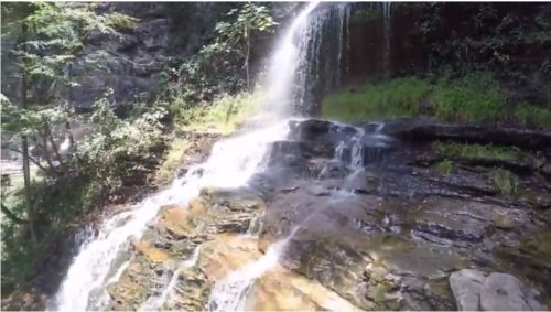 A cascading waterfall flows over rocky terrain, surrounded by lush greenery and trees.