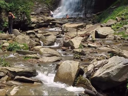 A serene waterfall cascades over rocks, with people enjoying the natural scenery by a flowing stream.