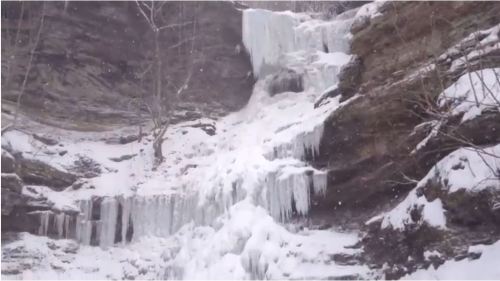 A frozen waterfall cascading down rocky cliffs, surrounded by snow-covered trees and a winter landscape.