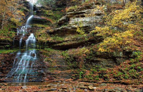 A serene waterfall cascading down rocky cliffs, surrounded by autumn foliage and a tranquil stream below.