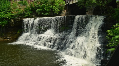 A serene waterfall cascading over rocks into a calm river, surrounded by lush green trees and a bridge above.