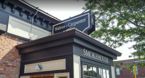 Sign for "Walrus + Carpenter Smokehouse" on a building with a brick facade and greenery above.