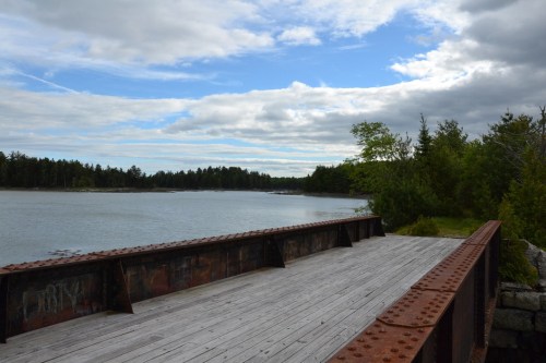 A wooden dock extends over calm water, surrounded by trees and a cloudy blue sky.