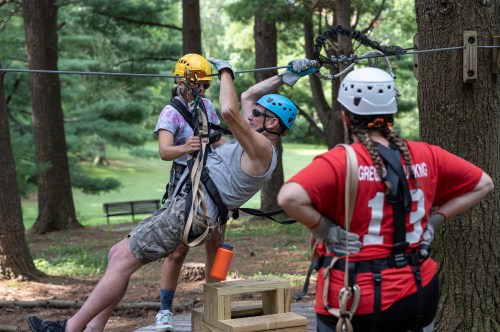 A man zip-lining with a child, while a woman in a red shirt watches, surrounded by trees in a park setting.