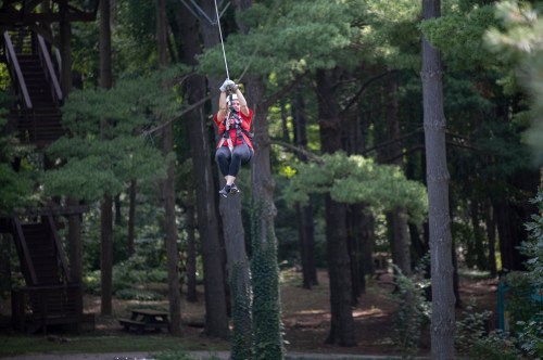A person in a red shirt zip-lining through a forested area, surrounded by tall trees and greenery.