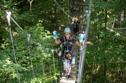 A person in a helmet and gloves smiles and waves while crossing a suspension bridge in a forested area.