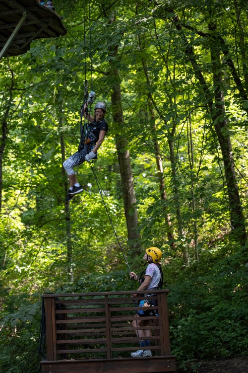 A person in a helmet rappels down from a platform while another person in a helmet assists from below in a wooded area.