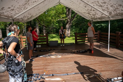 A group of people stands under a tent, preparing for an outdoor activity with harnesses and equipment on the ground.
